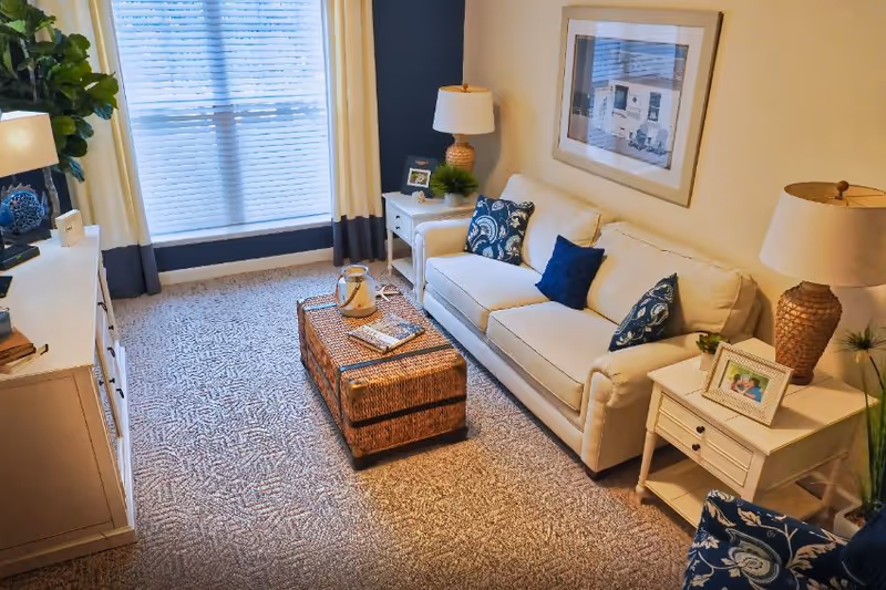 A cozy living room with a white sofa adorned with blue and patterned cushions, two side tables each with a lamp and decorative items, a wicker coffee table with a tray and books, a large window with white and navy curtains, a patterned carpet, and a framed picture on the wall.