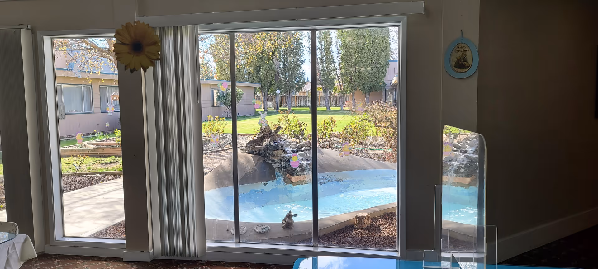 View through large sliding glass doors showing a landscaped outdoor courtyard with a small water feature, green grass, trees, and surrounding buildings. Inside, part of a table and a clear protective barrier are visible near the glass doors.
