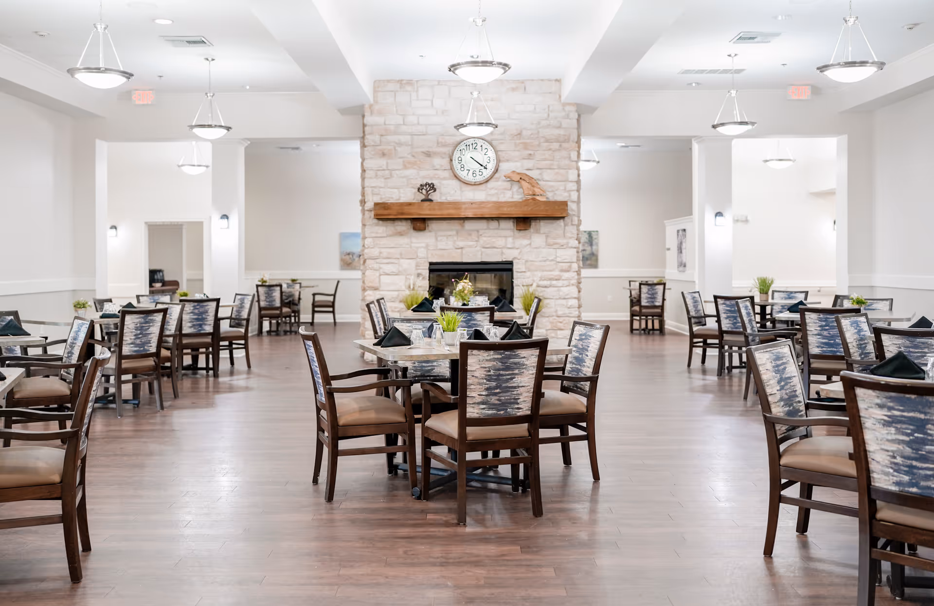 Large, bright dining room with multiple set tables and chairs centered around a stone fireplace and clock.