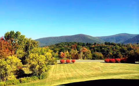 A scenic outdoor view featuring a large grassy lawn with neatly trimmed bushes and trees displaying autumn colors. In the background, there are forested hills and mountains under a clear blue sky.