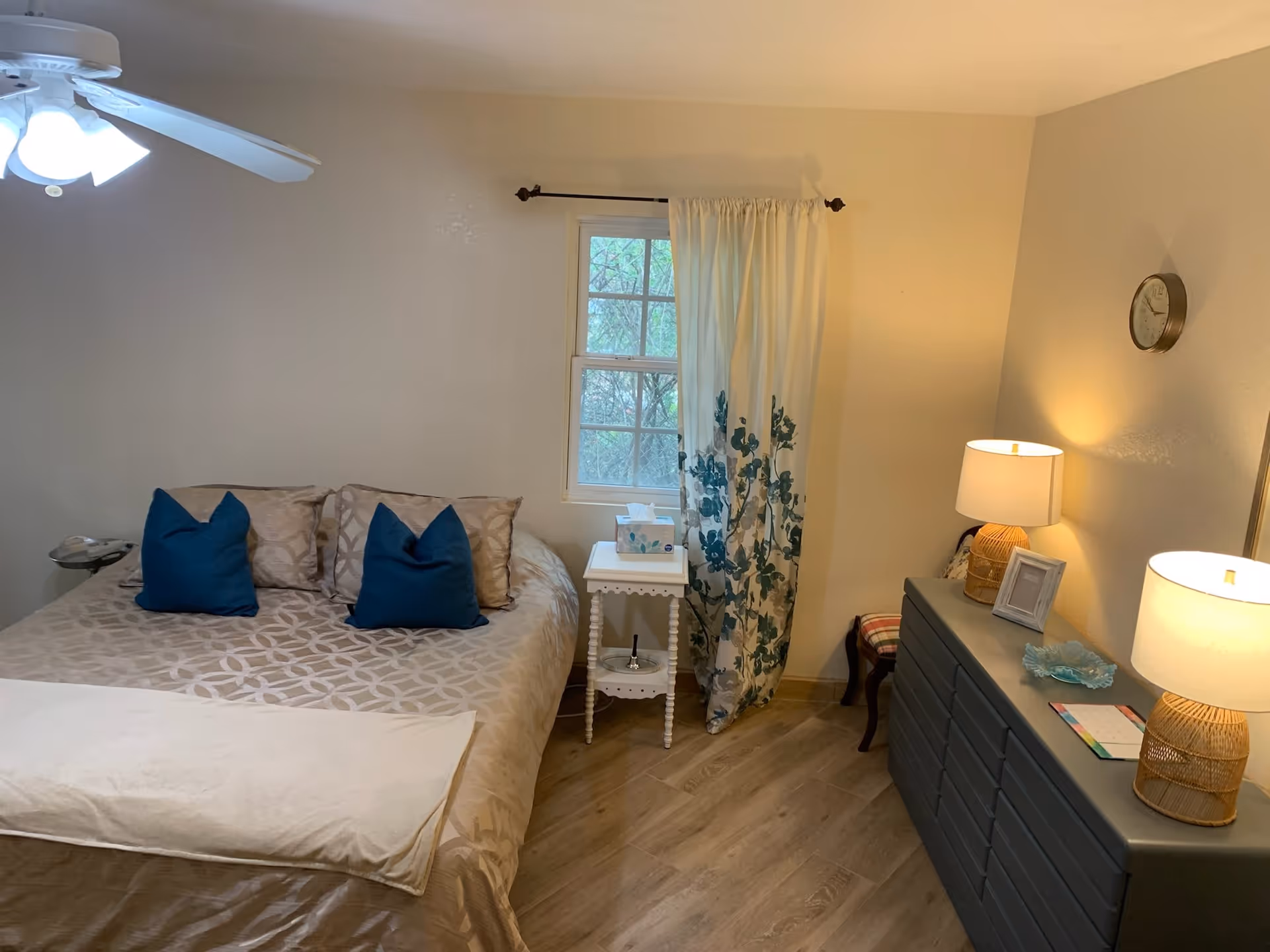 A cozy bedroom with a bed featuring beige patterned bedding and two blue pillows. There is a ceiling fan with lights above the bed. Next to the bed is a small white side table with a box of tissues. A window with white curtains decorated with blue floral patterns is behind the side table. On the right side of the room, there is a dark dresser with two lamps, a small framed picture, a decorative dish, and a calendar. A small chair with a plaid cushion is partially visible beside the dresser. The room has light-colored walls and wood flooring.