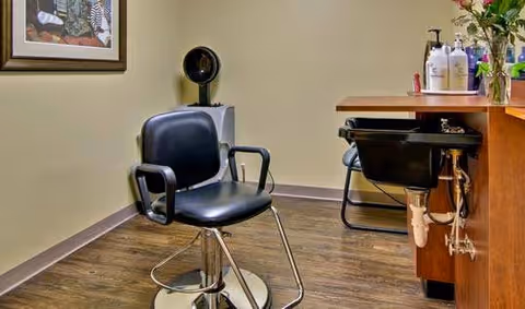 Interior view of a salon area with a black salon chair in the foreground, a hair washing station with a black sink and chair, and various hair care products on a wooden counter. The floor is wood and a framed picture hangs on the wall.