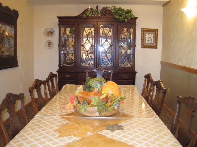 A traditional dining room with a long table covered by a beige and white checkered tablecloth. The table is decorated with a centerpiece featuring autumn-themed items such as a pumpkin, leaves, and flowers. Surrounding the table are wooden chairs with carved backs. Against the far wall is a large wooden china cabinet with glass doors displaying dishes and decorative items. The walls have framed artwork and decorative plates, and there is a wall sconce light on the right side.