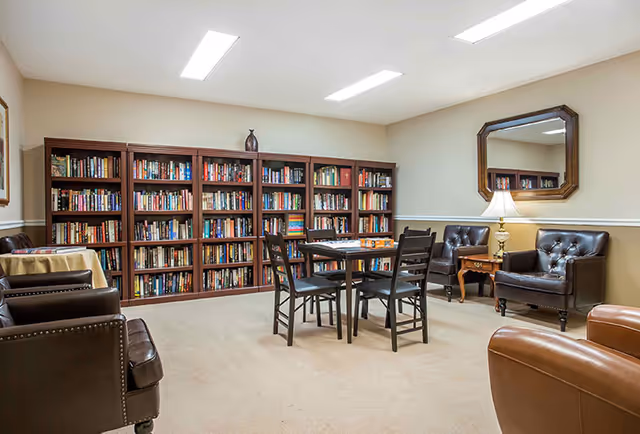 Well-lit common room with bookshelves lining the wall, a central table and chairs, and leather armchairs.