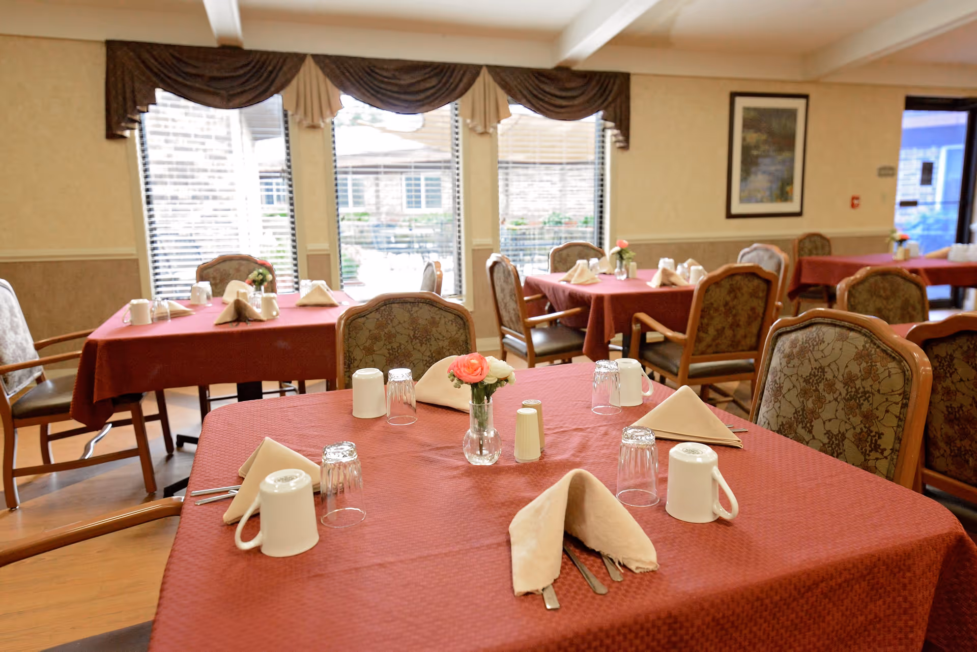 Dining room with multiple tables covered in red tablecloths, each set with beige napkins, white mugs, upside-down glasses, and small vases with flowers. The room has large windows with brown valances and a framed picture on the wall.