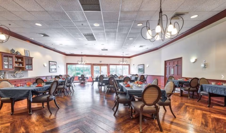 A spacious dining room with multiple tables covered in dark tablecloths, each set with napkins and small centerpieces. The room features wooden floors, large windows letting in natural light, chandeliers hanging from the ceiling, and framed artwork on the walls.
