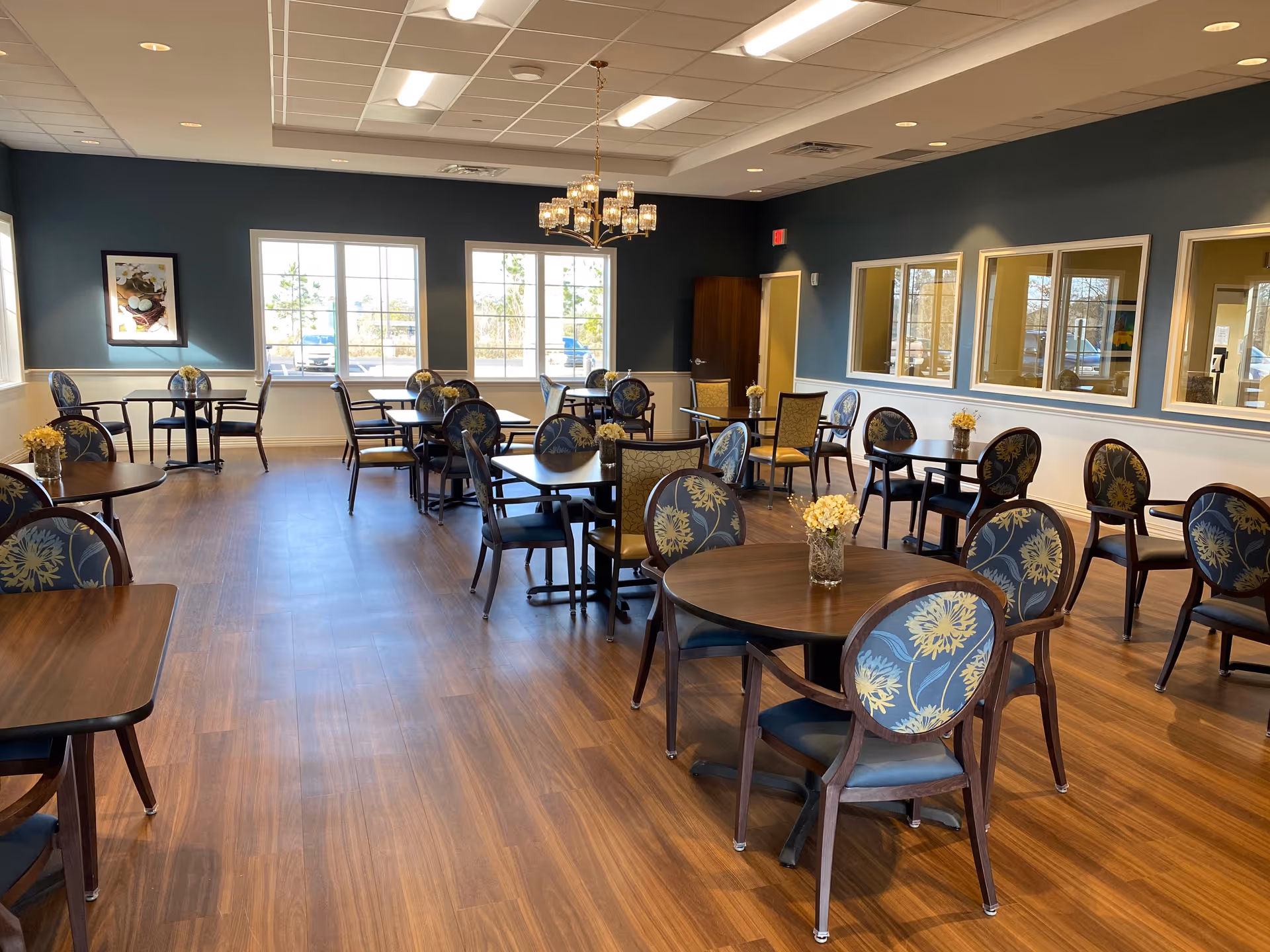 Spacious dining room with round wooden tables and floral-upholstered chairs, wood flooring, windows and a chandelier.
