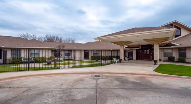 Exterior view of Rowlett Health & Rehabilitation Center showing a single-story building with a covered entrance, a driveway, and a fenced courtyard with green grass and benches under a cloudy sky.
