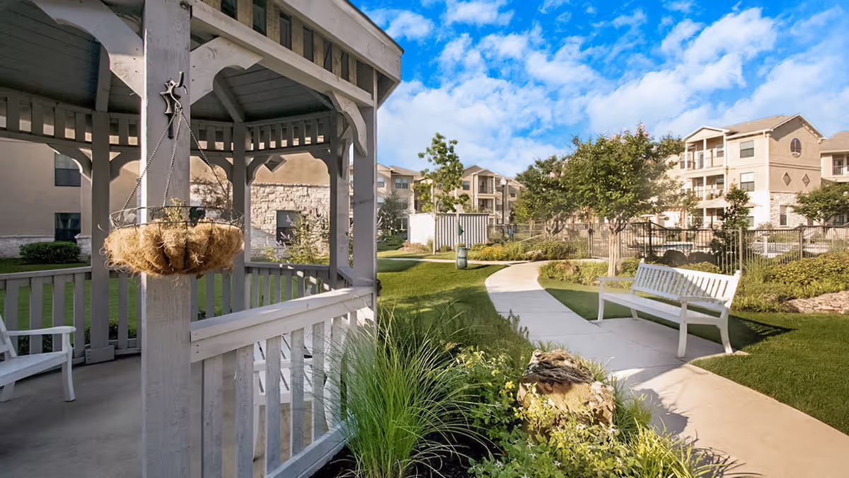 Outdoor garden area at Chateau McKinney featuring a white gazebo with hanging basket, white benches along a curved concrete pathway, green grass, trees, and residential buildings in the background under a partly cloudy blue sky.