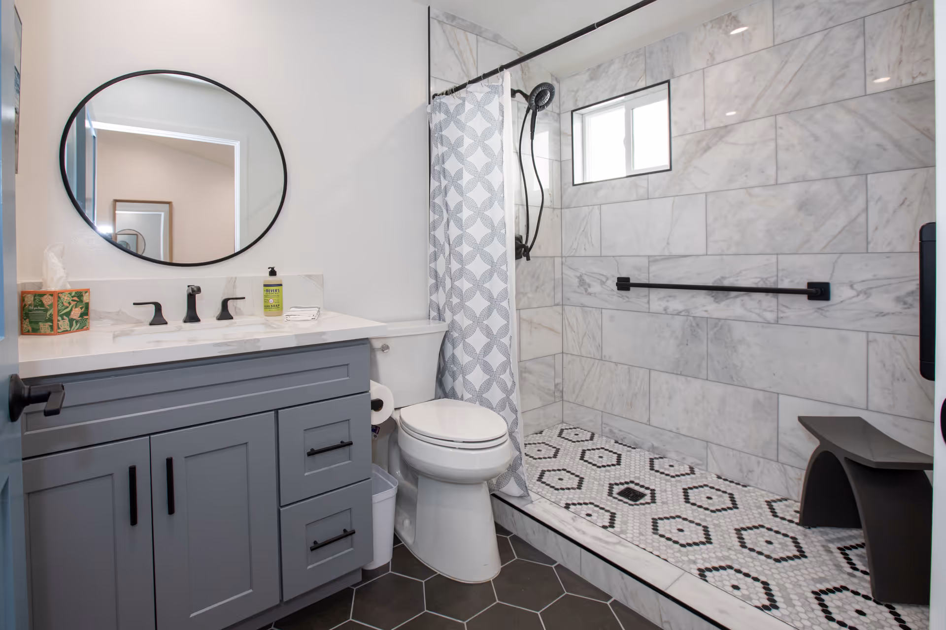 A modern bathroom featuring a gray vanity with black handles, a white countertop with a soap dispenser and tissue box, a round mirror above the sink, a white toilet, and a walk-in shower with patterned tile flooring, marble wall tiles, a black showerhead, a black grab bar, and a small black bench. The shower has a window and a patterned shower curtain.