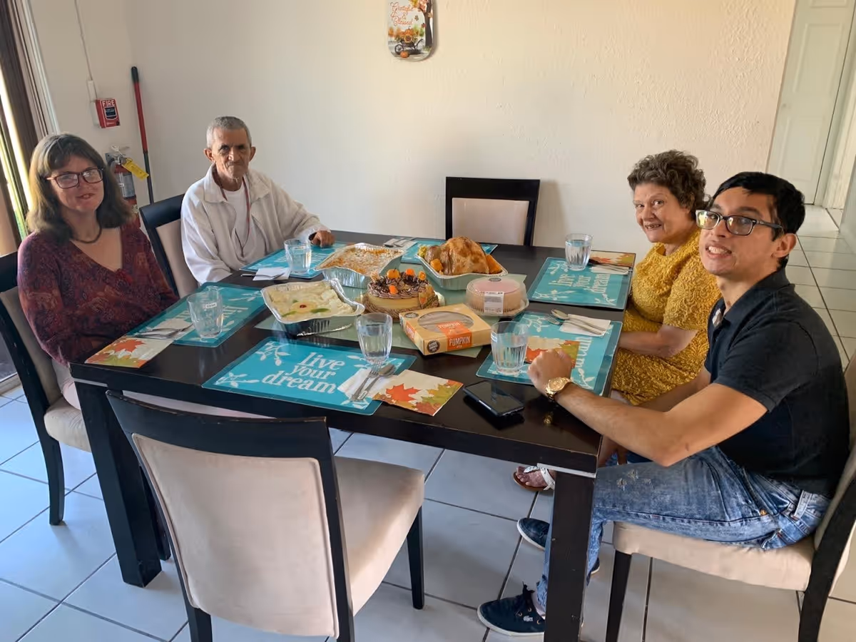 Four people sitting around a dining table set with placemats, glasses of water, and various dishes including a cake, a roasted chicken, and other food items in a well-lit room.