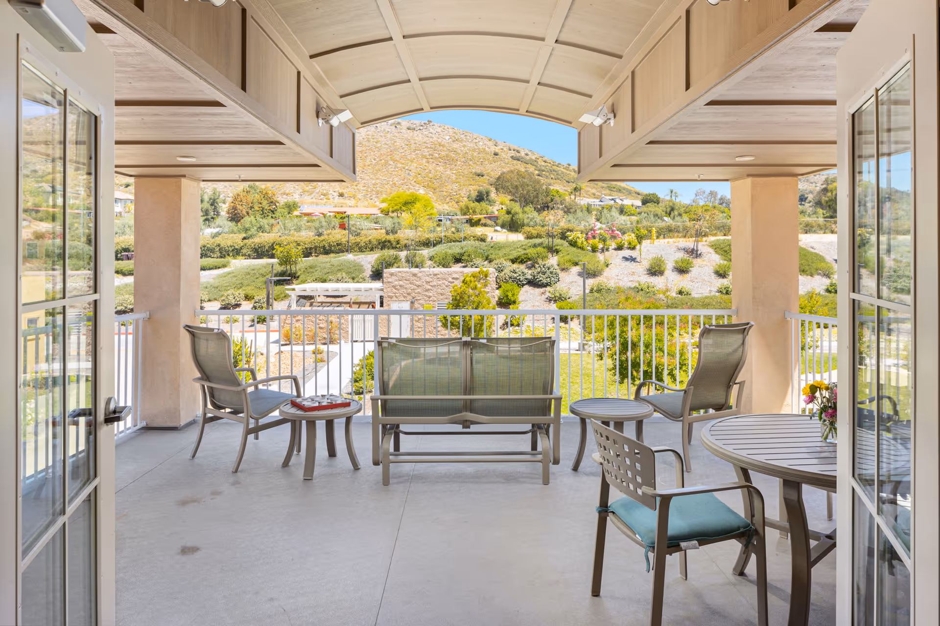 Covered outdoor patio area with metal chairs, tables, and a bench overlooking a landscaped garden and hills in the background under a clear blue sky.