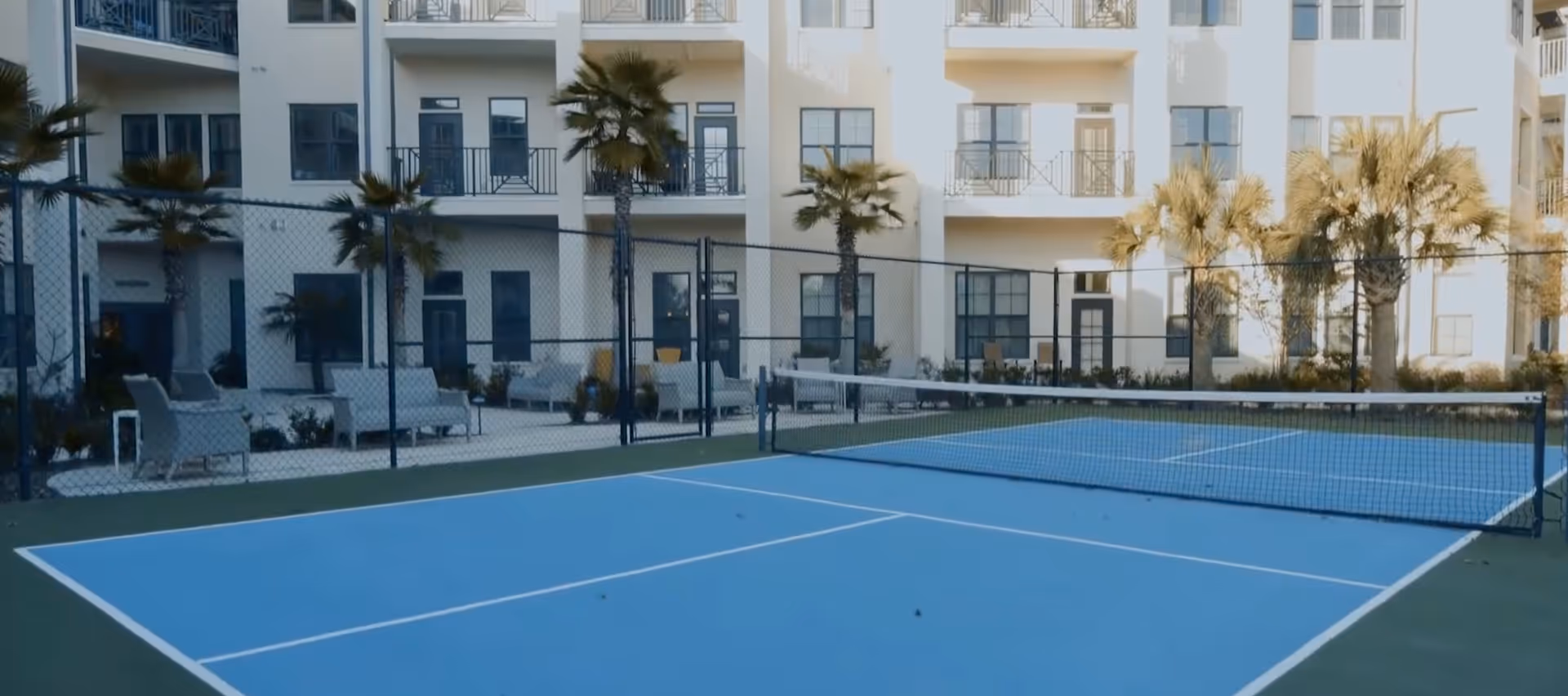 Outdoor tennis court with a blue playing surface and white lines, surrounded by a black chain-link fence. Behind the court, there are several palm trees and a multi-story building with balconies and large windows. Outdoor seating with chairs and small tables is visible near the building.