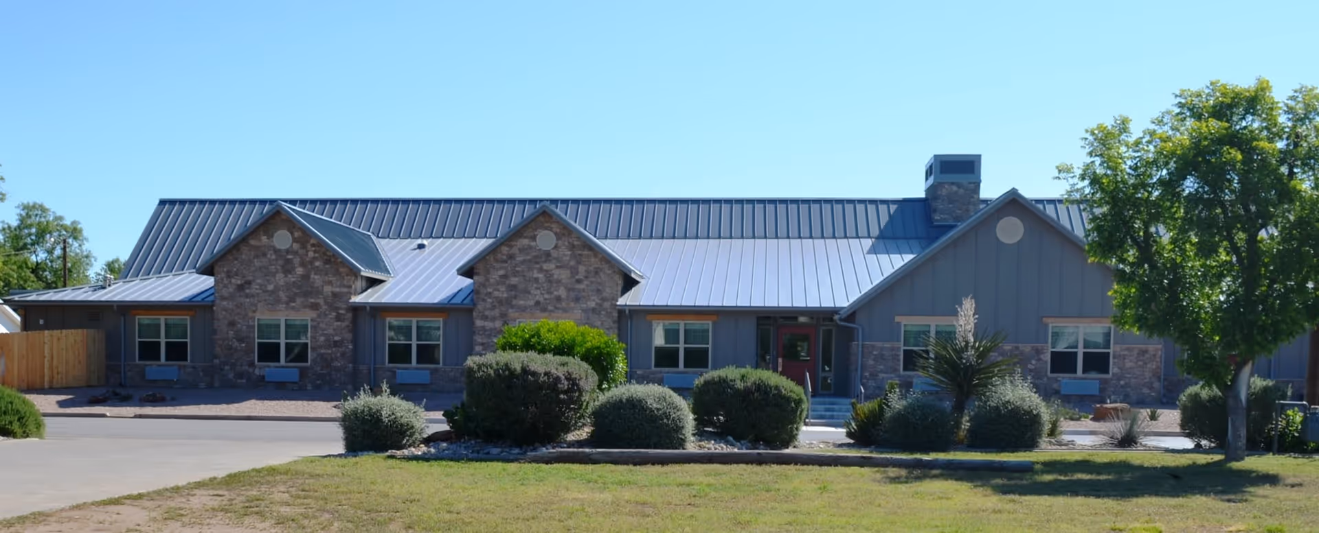 Front exterior view of a single-story building with a metal roof, stone and siding facade, several windows, a chimney, and landscaped bushes and trees in front under a clear blue sky.