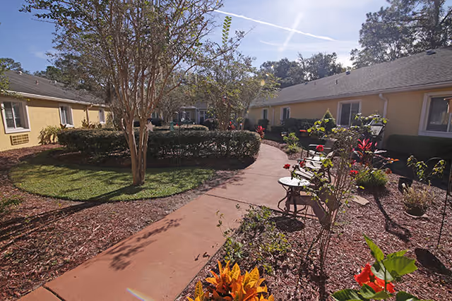 Sunny landscaped courtyard with a paved walkway, benches, flowering plants, and single-story yellow buildings surrounding it.