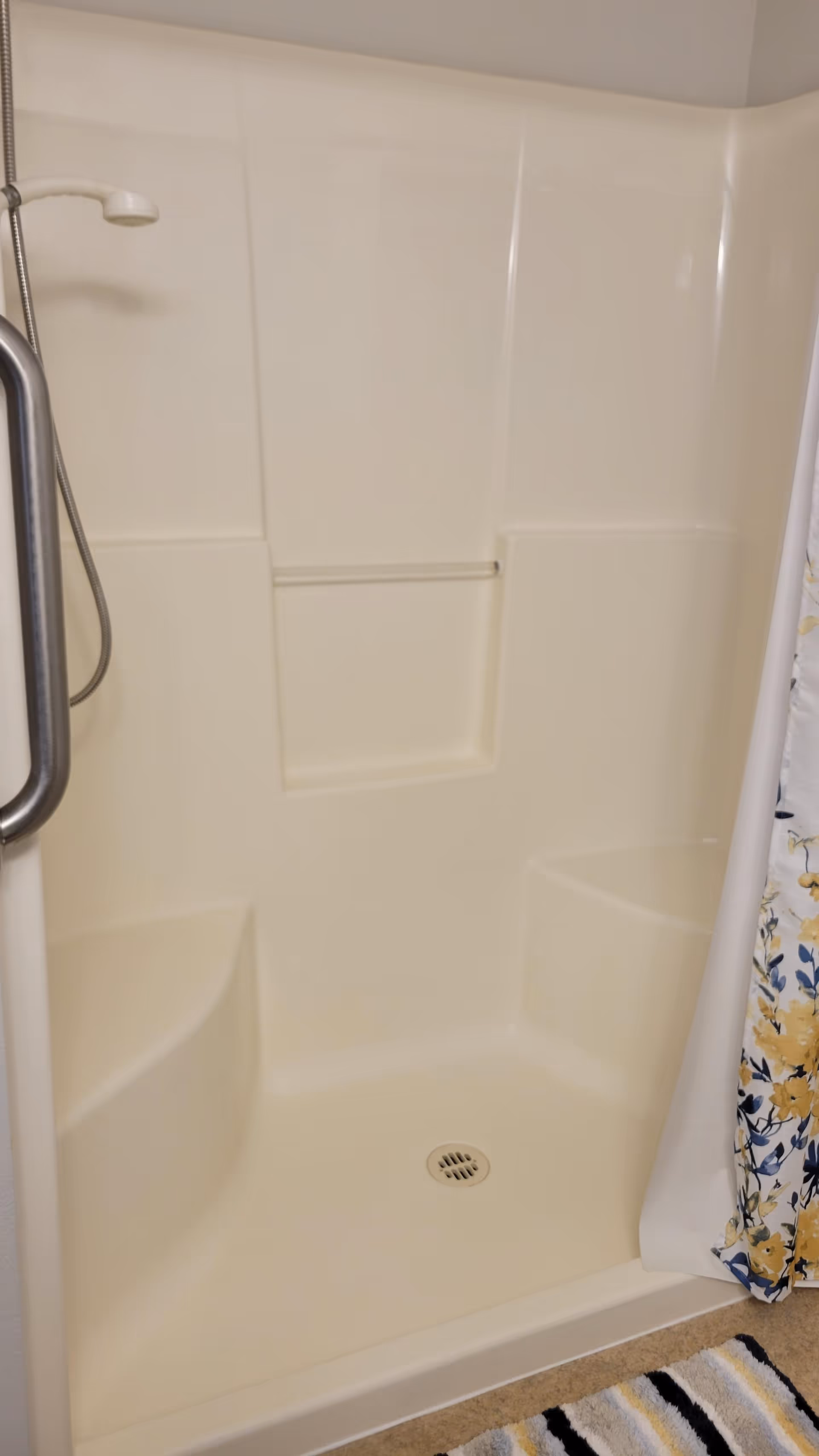 A beige-colored shower stall with built-in corner seats on both sides and a small recessed shelf in the middle. There is a handheld showerhead attached to a metal hose on the left side and a metal grab bar. A floral shower curtain with yellow and blue flowers is partially visible on the right side, and a striped bath mat is on the floor outside the shower.