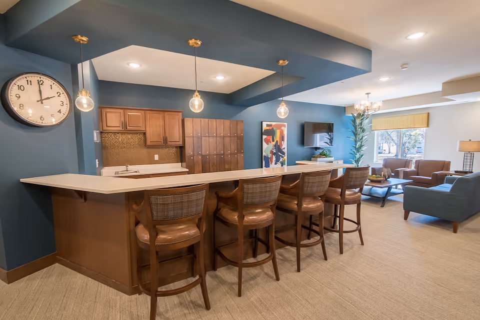 A spacious common area with a long counter and five wooden bar stools with leather seats. Behind the counter is a small kitchenette with wooden cabinets and a sink. The walls are painted blue, and there is a large clock on the left wall. The room also features a seating area with armchairs, a coffee table, a flat-screen TV mounted on the wall, and a large window letting in natural light. Three pendant lights hang above the counter.