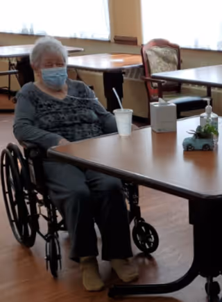 An elderly woman wearing a face mask sitting in a wheelchair at a table in a communal dining or activity room. The table has a white disposable cup with a straw, a tissue box, and a small decorative plant. The room has wooden floors and large windows with curtains, and there are other tables and chairs in the background.