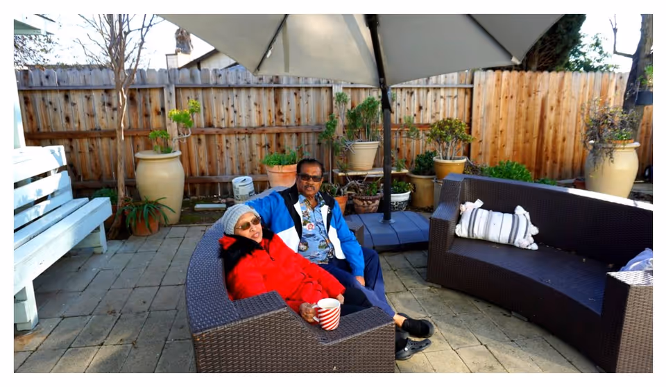 Two people sitting together on curved outdoor wicker patio sofas under a large umbrella on a paved patio with potted plants and a wooden fence behind them.