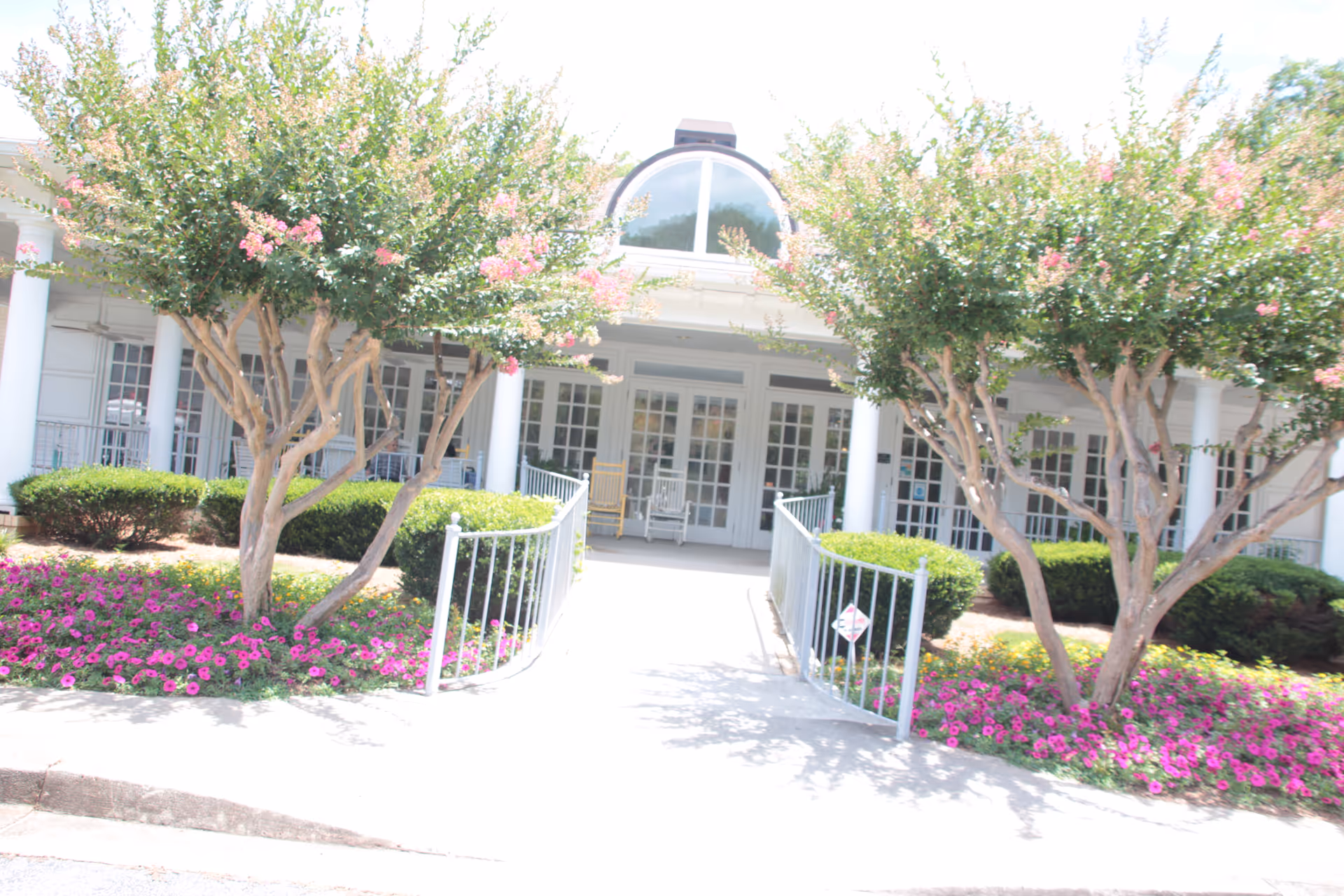 Front entrance of a senior living building with a ramp, white columns, double glass doors, and flowering trees and beds.