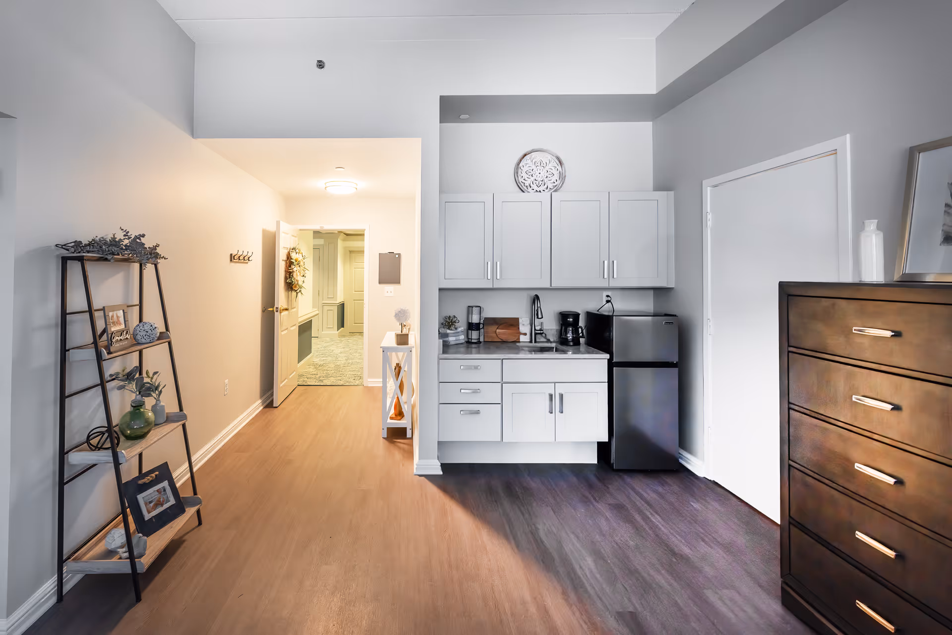 Interior view of a senior living facility room featuring a small kitchenette with white cabinets, a sink, coffee maker, and mini refrigerator. To the right is a dark wooden dresser with a framed picture and a decorative vase on top. On the left side, there is a black metal and wood shelving unit with various decorative items. The hallway beyond the kitchenette is visible, leading to other rooms with light-colored walls and flooring.