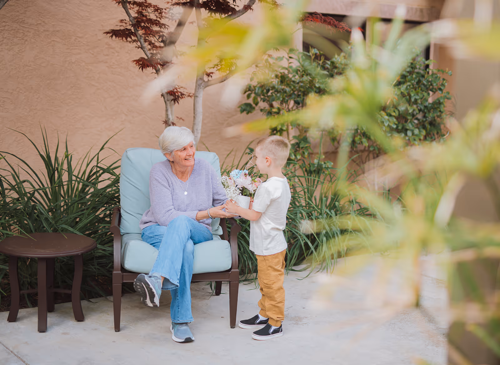 An elderly woman sitting on a cushioned outdoor chair smiling at a young boy who is standing and holding a potted plant with flowers. They are in a garden-like outdoor area with green plants and a small round table nearby.