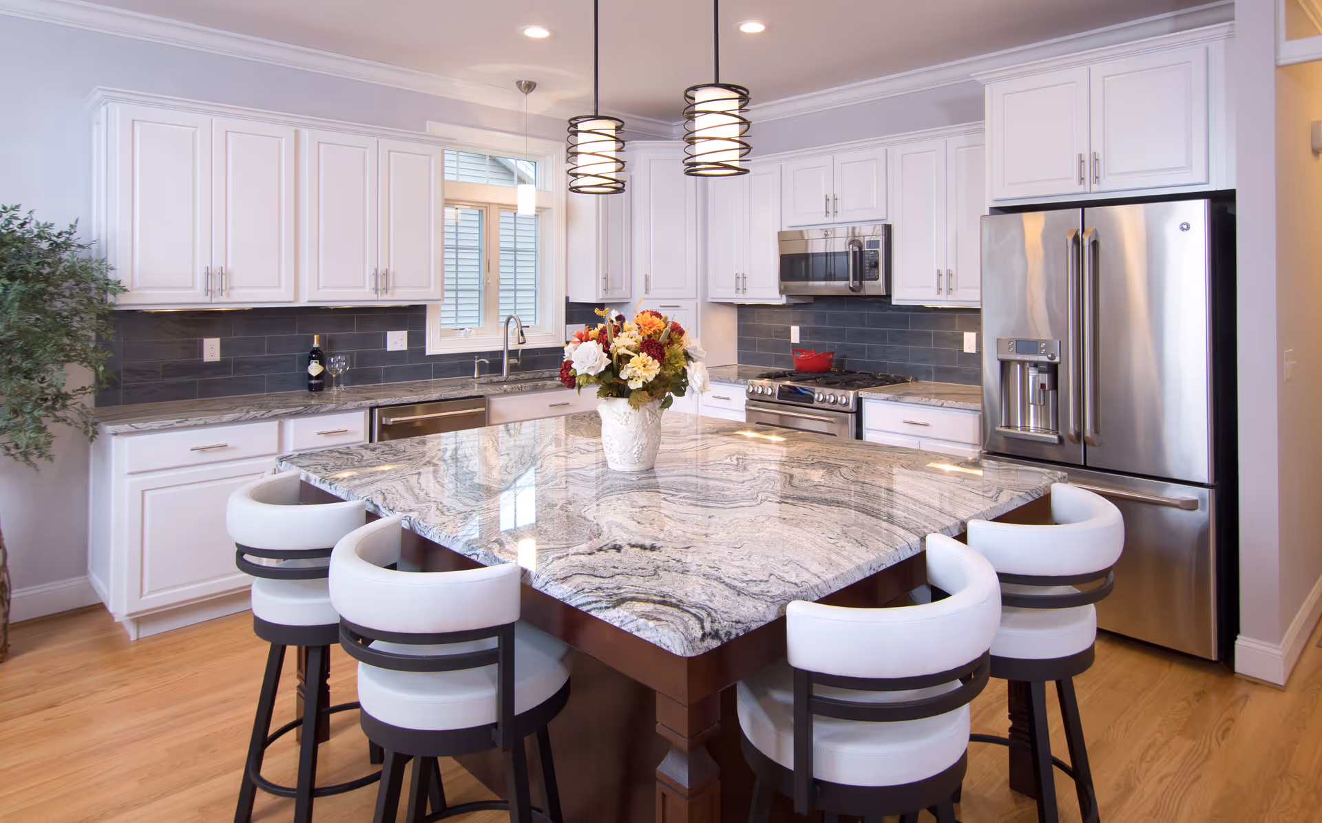 Bright modern kitchen with a large marble island surrounded by white cushioned bar stools, white cabinets, stainless steel appliances and pendant lights.