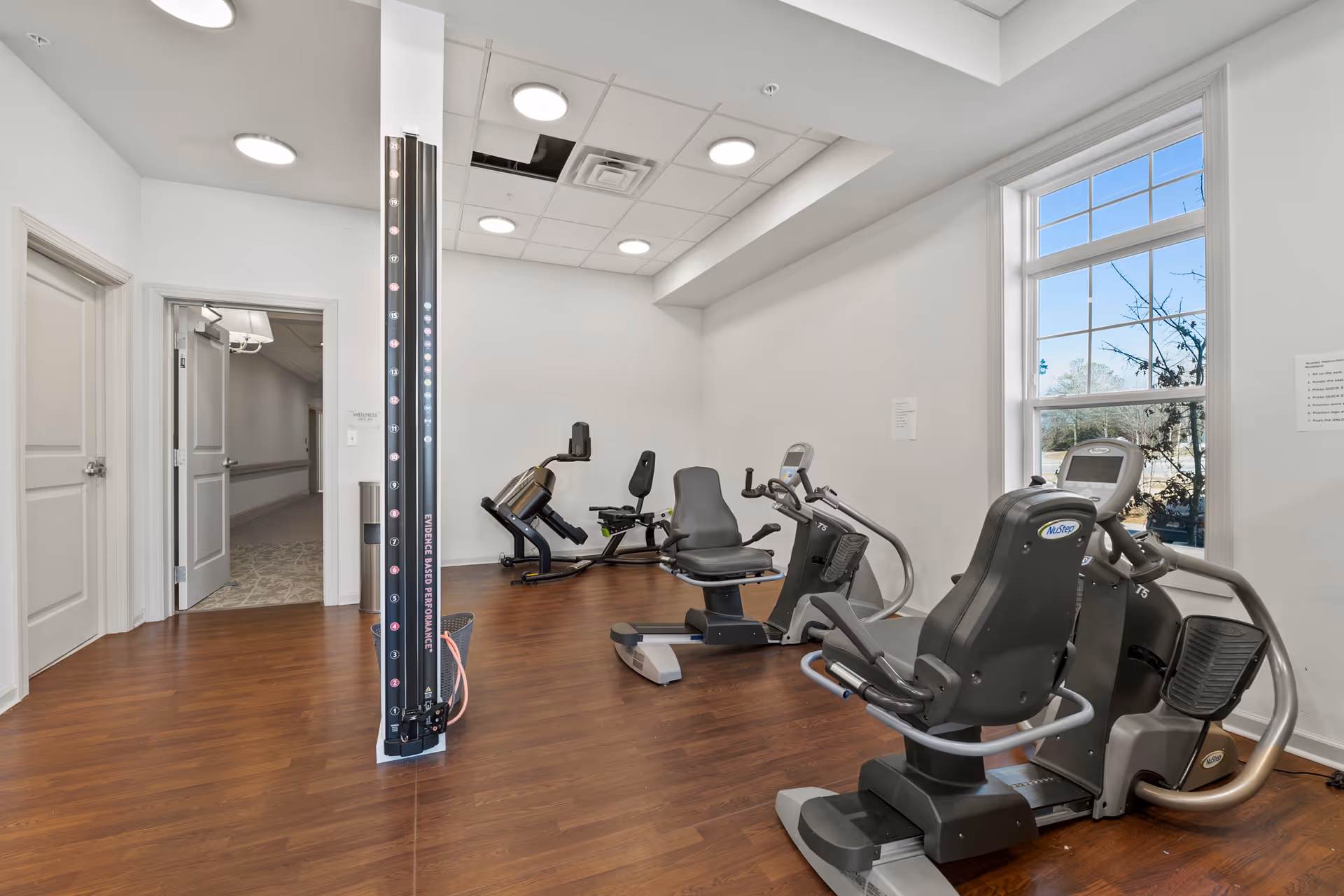 A small fitness room with exercise equipment including recumbent bikes and a leg press machine. The room has wooden flooring, white walls, a large window letting in natural light, and recessed ceiling lights. There is an open door leading to a hallway.