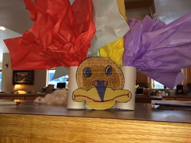 Handmade turkey craft made from toilet paper rolls and colored tissue paper sitting on a wooden countertop in a communal room.