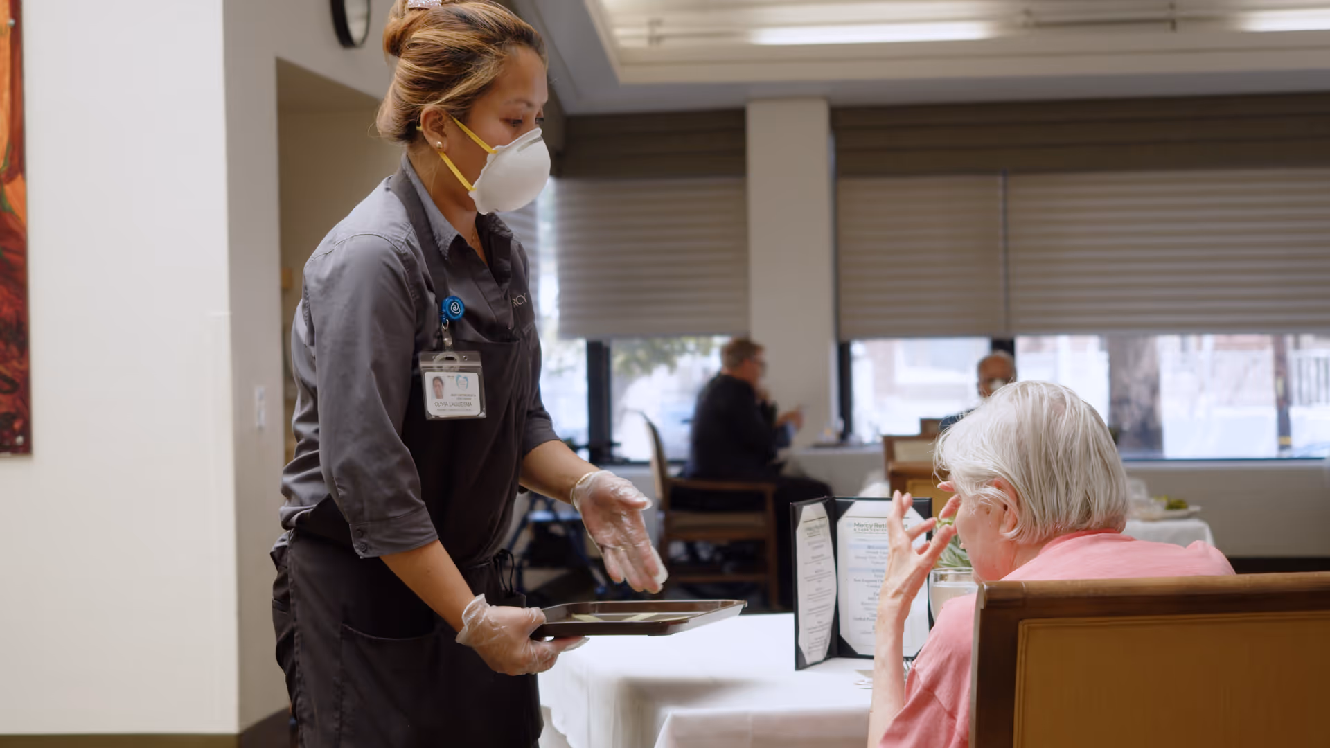 A masked staff member wearing gloves serves a tray to an elderly woman seated at a dining table in a communal dining area.