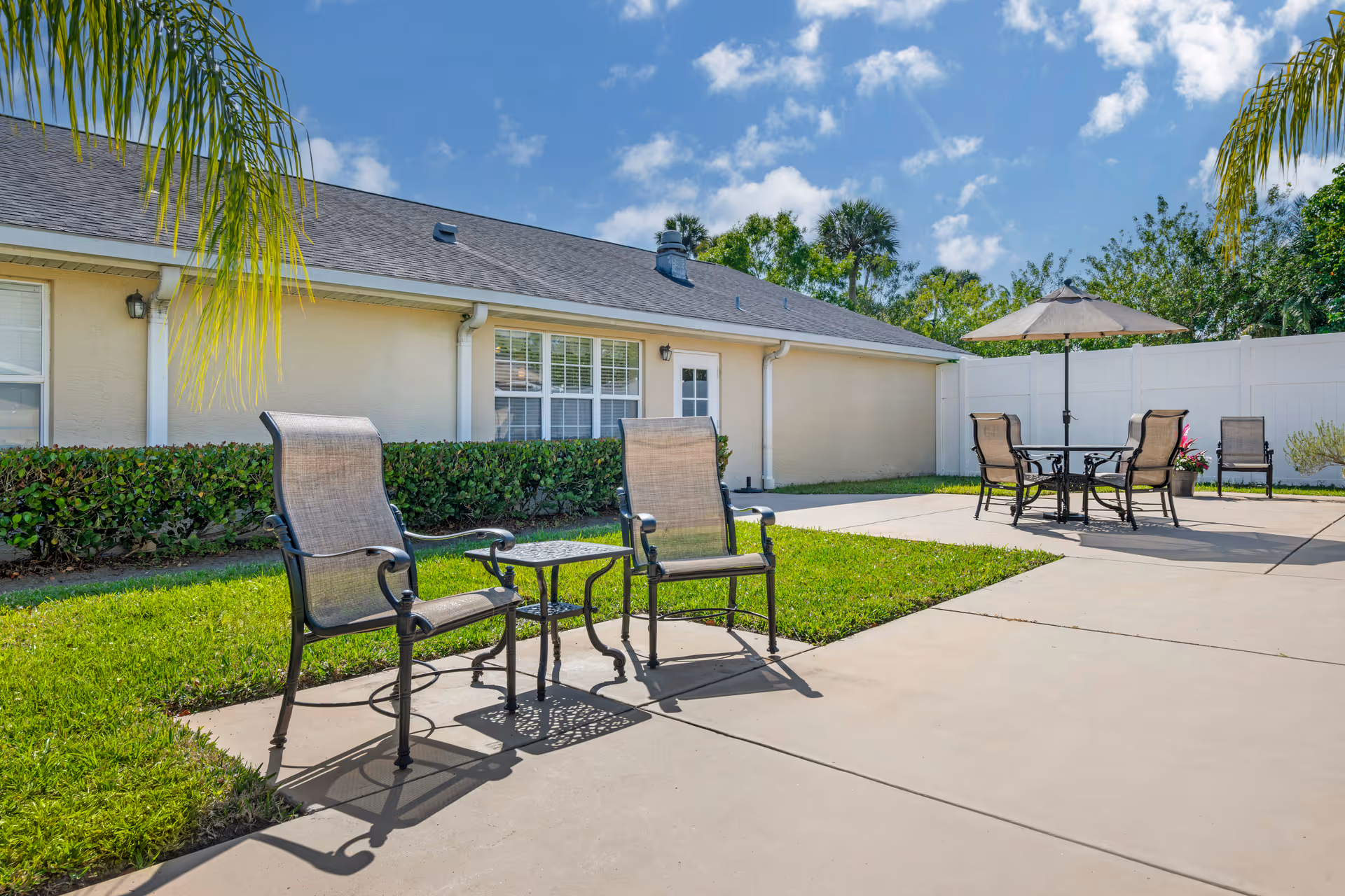 Outdoor patio area at Brookdale Vero Beach South Memory Care featuring metal chairs with mesh seats and backs, a small metal table, a round table with an umbrella, and additional chairs. The patio is surrounded by green grass, bushes, a white privacy fence, and a beige building under a blue sky with scattered clouds.