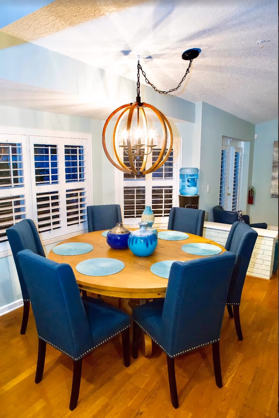 Round wooden dining table set with six blue upholstered chairs, blue placemats and decorative vases beneath a round hanging chandelier in a bright dining area.
