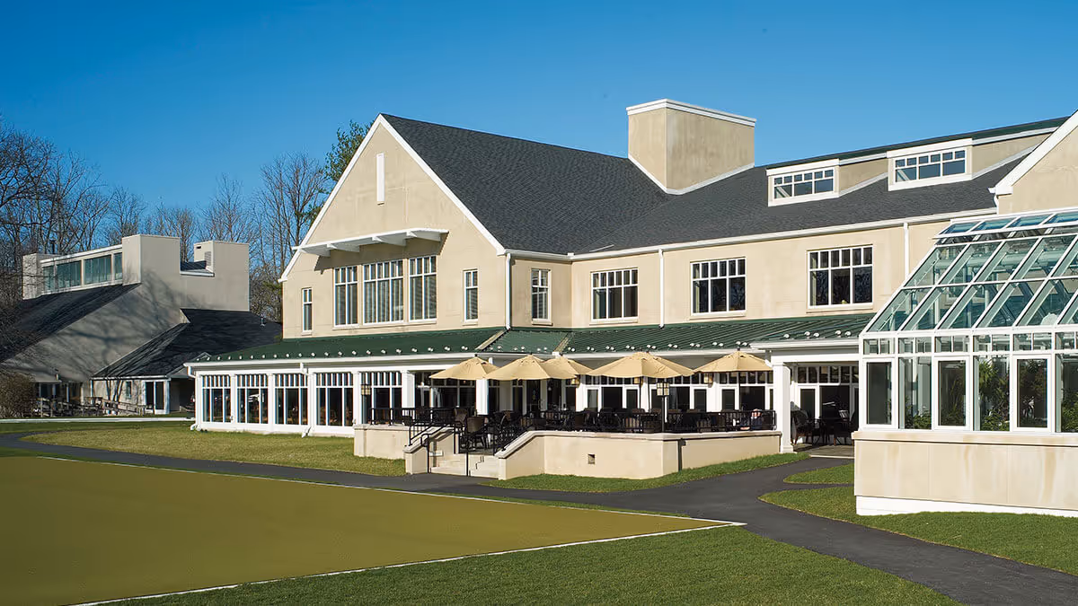 Front exterior of a beige senior living building with a patio, umbrellas, and a lawn.