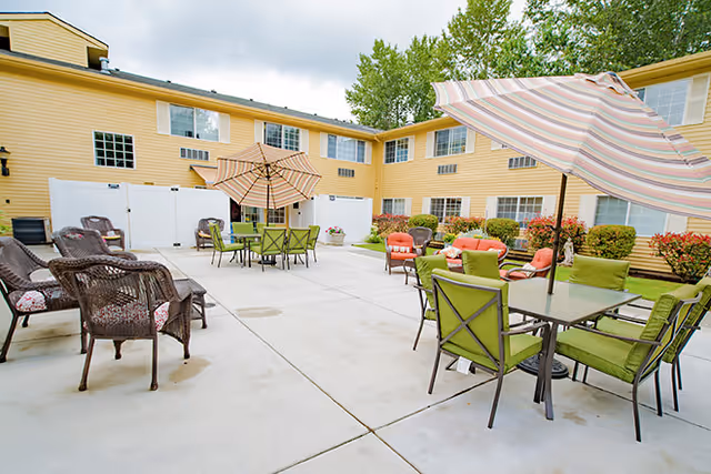 Outdoor courtyard area of a senior living facility with multiple seating arrangements including green cushioned chairs around tables with umbrellas, and brown wicker chairs with cushions. The courtyard is surrounded by a two-story yellow building with windows and some greenery along the edges.