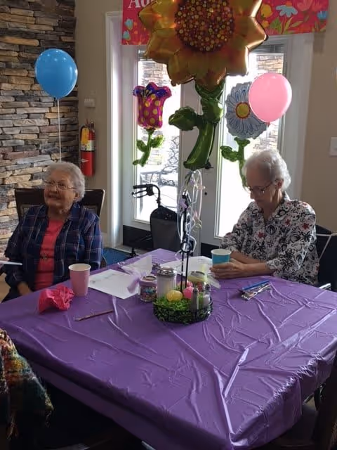 Two elderly women sitting at a table covered with a purple tablecloth in a room with stone wall accents. The table is decorated with colorful flower-shaped balloons and has cups, papers, and small containers on it. One woman is wearing a plaid shirt and smiling, while the other is wearing a floral shirt and looking down at a cup. There are blue and pink balloons tied to chairs in the background near a glass door.