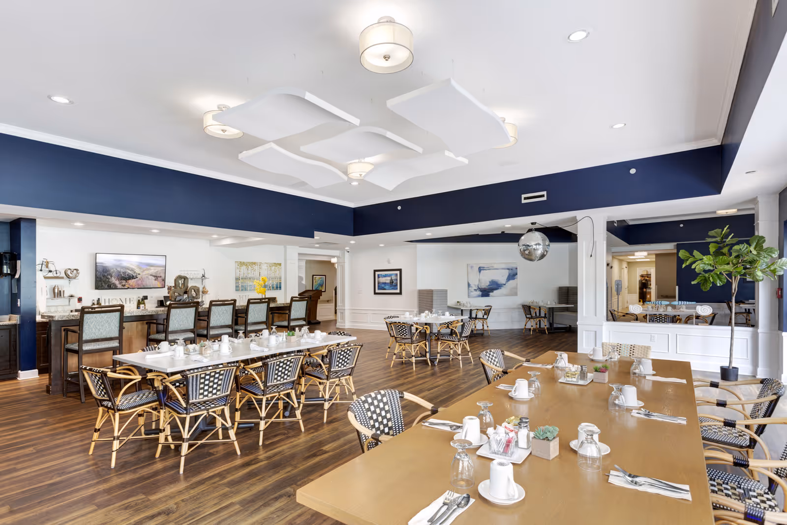 A spacious dining room with multiple tables set with white cups, glasses, and silverware. The room features wooden floors, a dark blue accent wall, and modern ceiling lights. There are several chairs with woven seats and backs around the tables, a bar area with high chairs, and decorative artwork on the walls. A large plant is placed near a window on the right side.