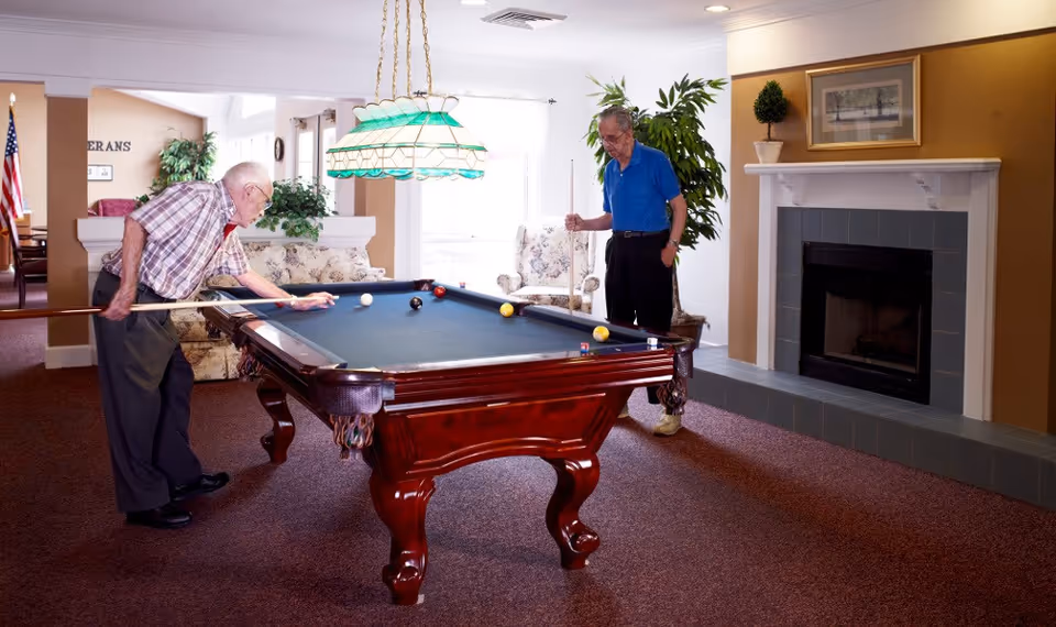 Two elderly men playing pool in a well-lit room with a fireplace, plants, and comfortable seating. One man is taking a shot while the other watches. The room has a carpeted floor and a hanging green and white stained glass light fixture above the pool table.