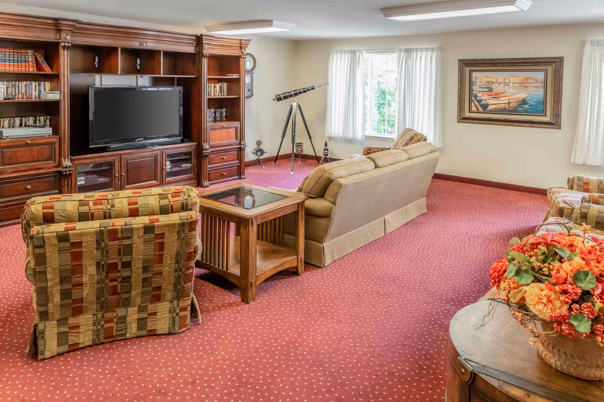 A cozy living room with a red patterned carpet, a beige sofa, two patterned armchairs, a wooden coffee table, and a large wooden entertainment center with a flat-screen TV and shelves filled with books and DVDs. There is a telescope near the window with white curtains, a framed painting of boats on the wall, and a decorative flower arrangement on a round wooden table in the foreground.