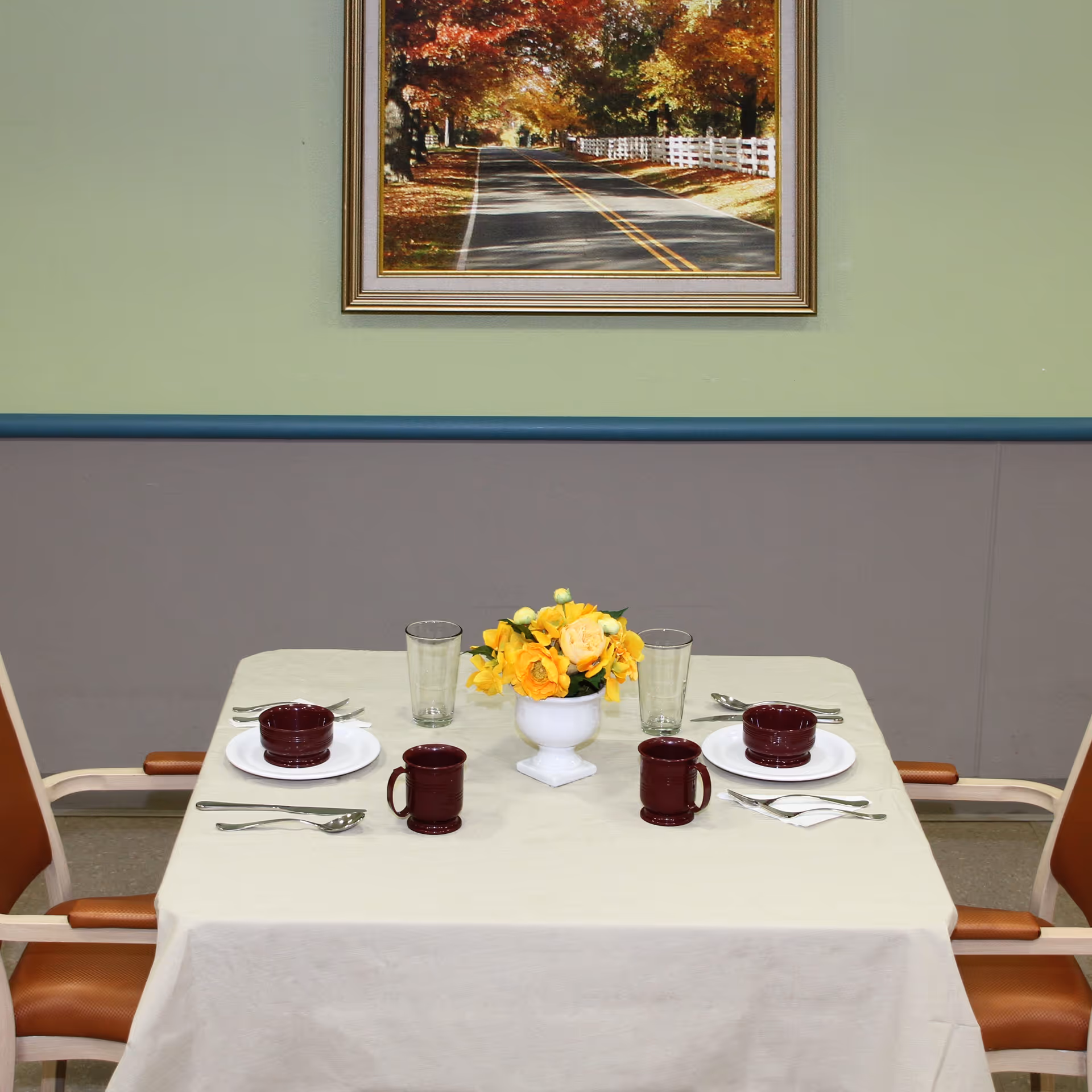A small dining table set for two with a beige tablecloth, two brown chairs, two place settings with white plates, maroon bowls and mugs, silverware, and two clear drinking glasses. A white vase with yellow flowers is centered on the table. A framed picture of a tree-lined road with autumn foliage hangs on the green wall behind the table.