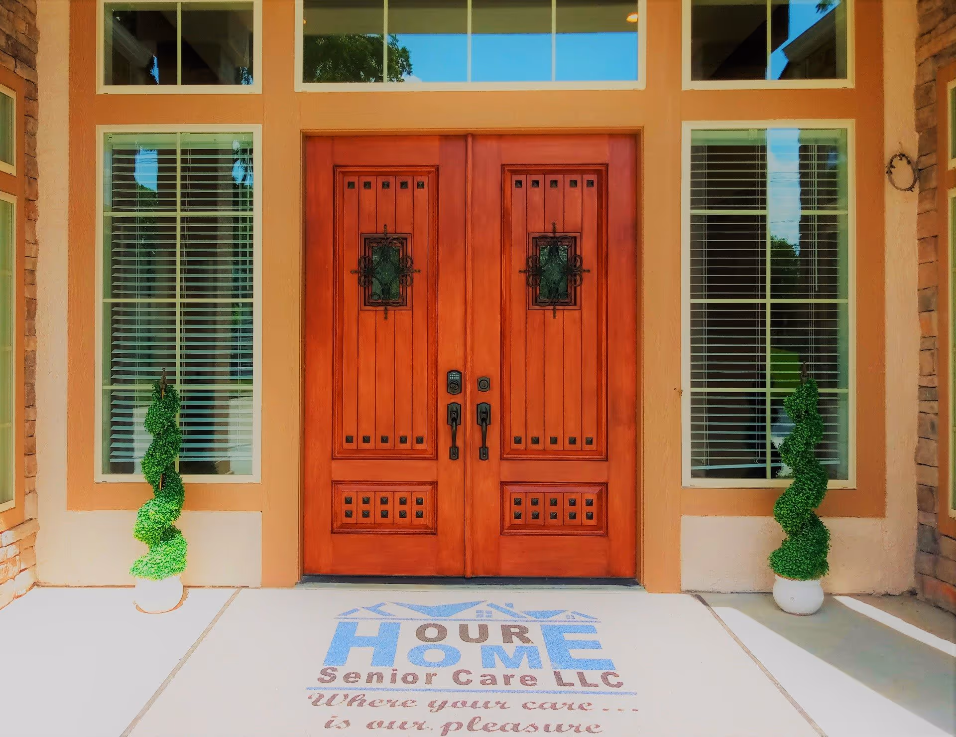 Front entrance of a building with double wooden doors featuring decorative glass panels. The doors are flanked by tall windows with white blinds. Two spiral-shaped topiary plants in white pots are placed symmetrically on either side of the entrance. A welcome mat in front of the doors displays the text 'Our Home Senior Care LLC Where your care... is our pleasure.'