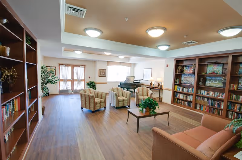 A spacious, well-lit common area with wooden floors and ceiling lights. The room features multiple bookshelves filled with books and decorative items, a piano in the back corner, several upholstered armchairs arranged around a wooden coffee table with a plant, and a sofa on the right side. Large windows and glass doors allow natural light to enter the room.