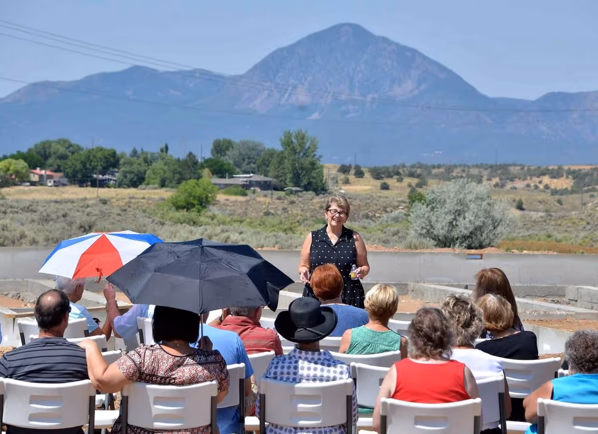 A group of elderly people seated outdoors on white chairs, some holding umbrellas for shade, listening to a woman standing and speaking in front of them with a mountainous landscape in the background.