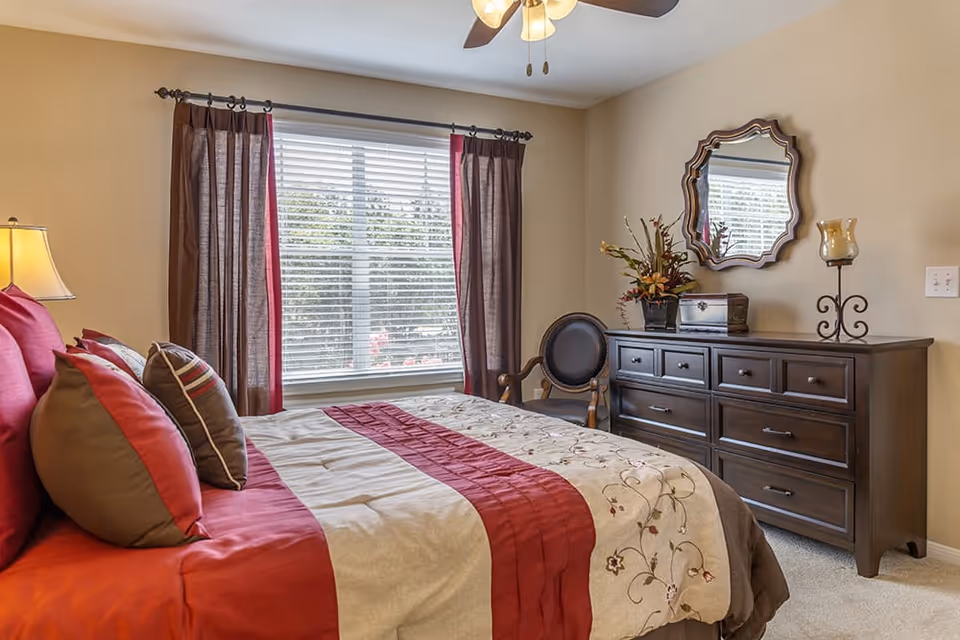 A cozy bedroom with a large bed featuring red and beige bedding and multiple pillows. There is a window with brown and red curtains letting in natural light. A dark wooden dresser with a decorative mirror above it, a chair, and a lamp on the left side complete the room.