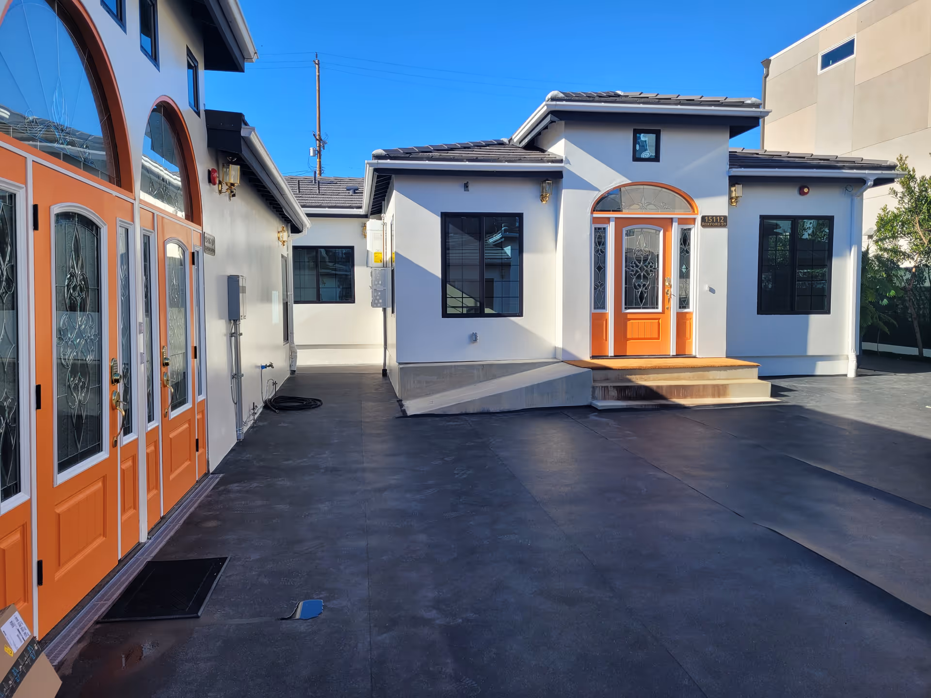 Exterior view of a building with white walls and orange doors and window frames. The building has a small set of stairs leading to an entrance door with decorative glass panels. The ground is paved with dark asphalt, and the sky is clear and blue.