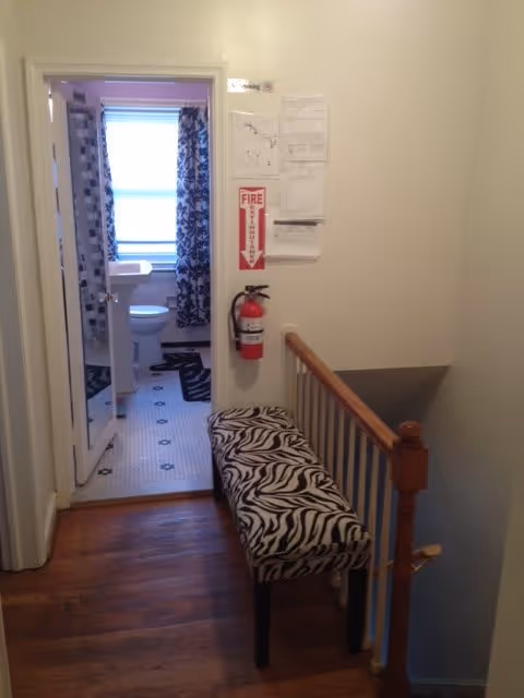 View of a hallway with wooden flooring leading to a bathroom. The bathroom has a pedestal sink, toilet, and a window with patterned curtains. A zebra-print bench is placed along the hallway wall next to a wooden railing. A fire extinguisher and some notices are mounted on the wall near the bathroom door.