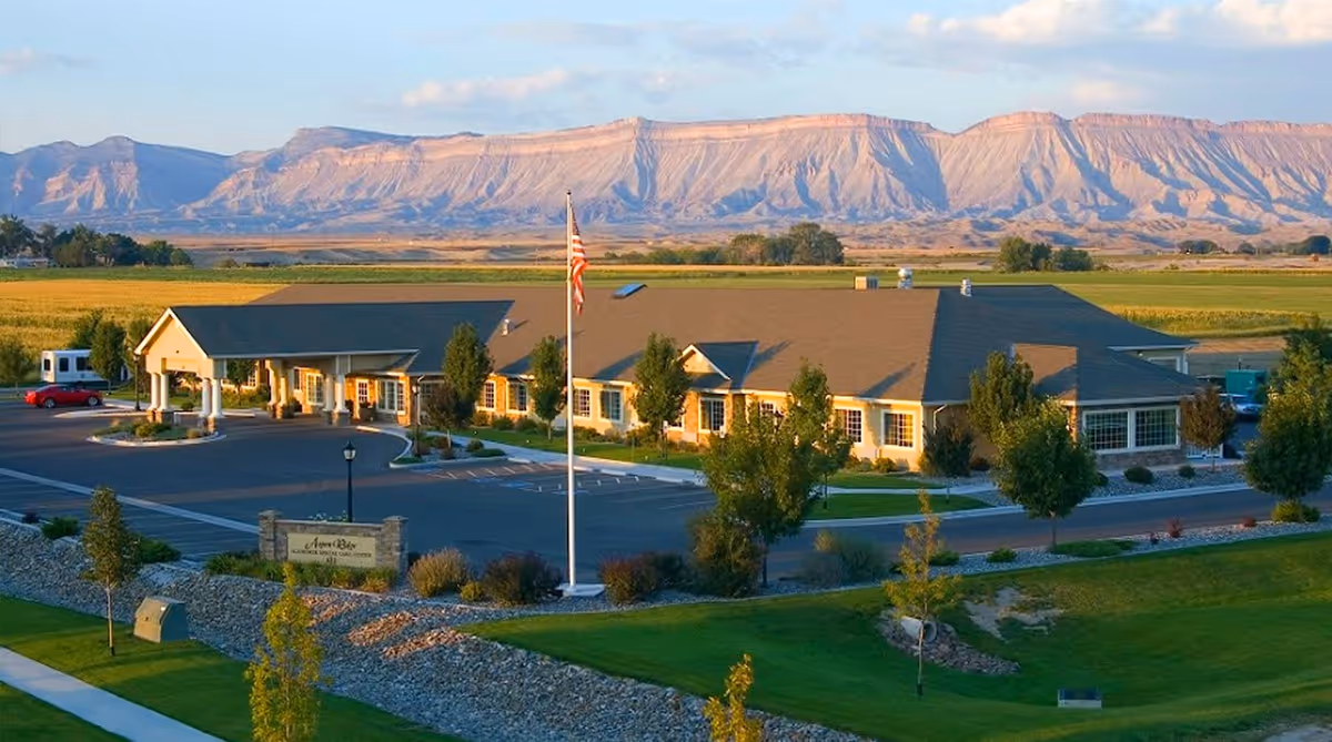 A single-story building with a large covered entrance and multiple windows, surrounded by a parking lot and landscaped greenery. An American flag is flying on a flagpole in front of the building. In the background, there are expansive fields and a range of flat-topped mountains under a partly cloudy sky.