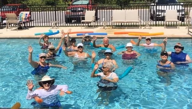 A group of elderly people in swimwear and hats participating in a water exercise class in an outdoor swimming pool, waving and smiling at the camera. Pool noodles float on the water surface, and poolside chairs and a fence are visible in the background.