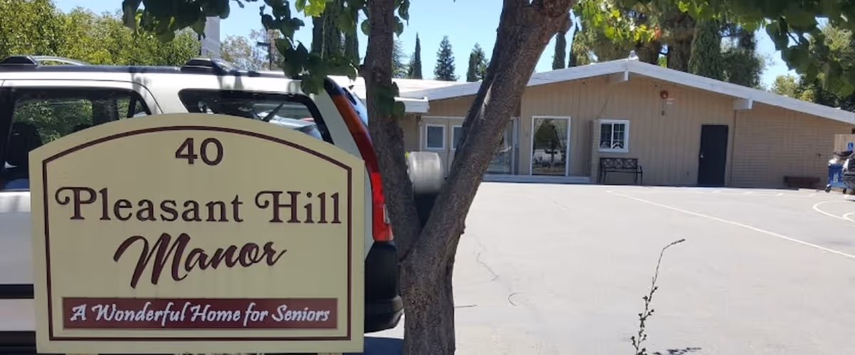 Exterior view of Pleasant Hill Manor senior living facility showing a beige building with windows and doors, a parking area with a white vehicle, and a sign in the foreground that reads '40 Pleasant Hill Manor A Wonderful Home for Seniors'. Trees and greenery are also visible.