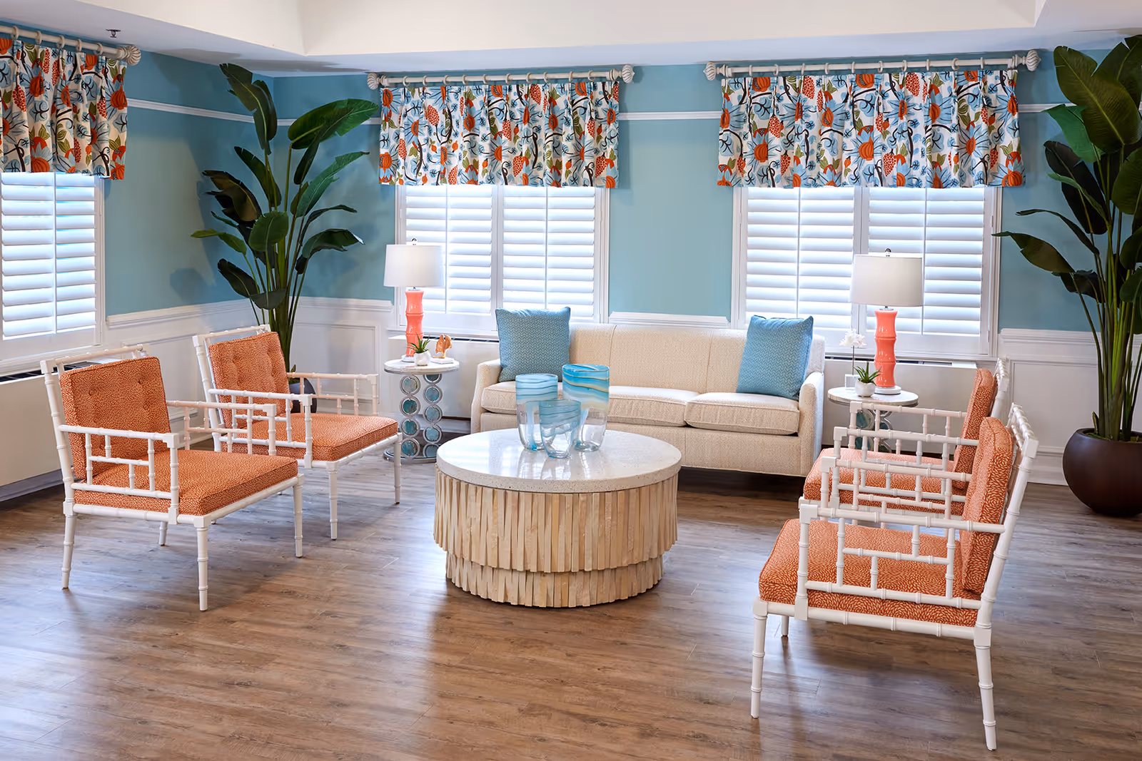 Bright common living room with coral-upholstered chairs, a beige sofa, round coffee table, potted plants, and teal walls with patterned valances.