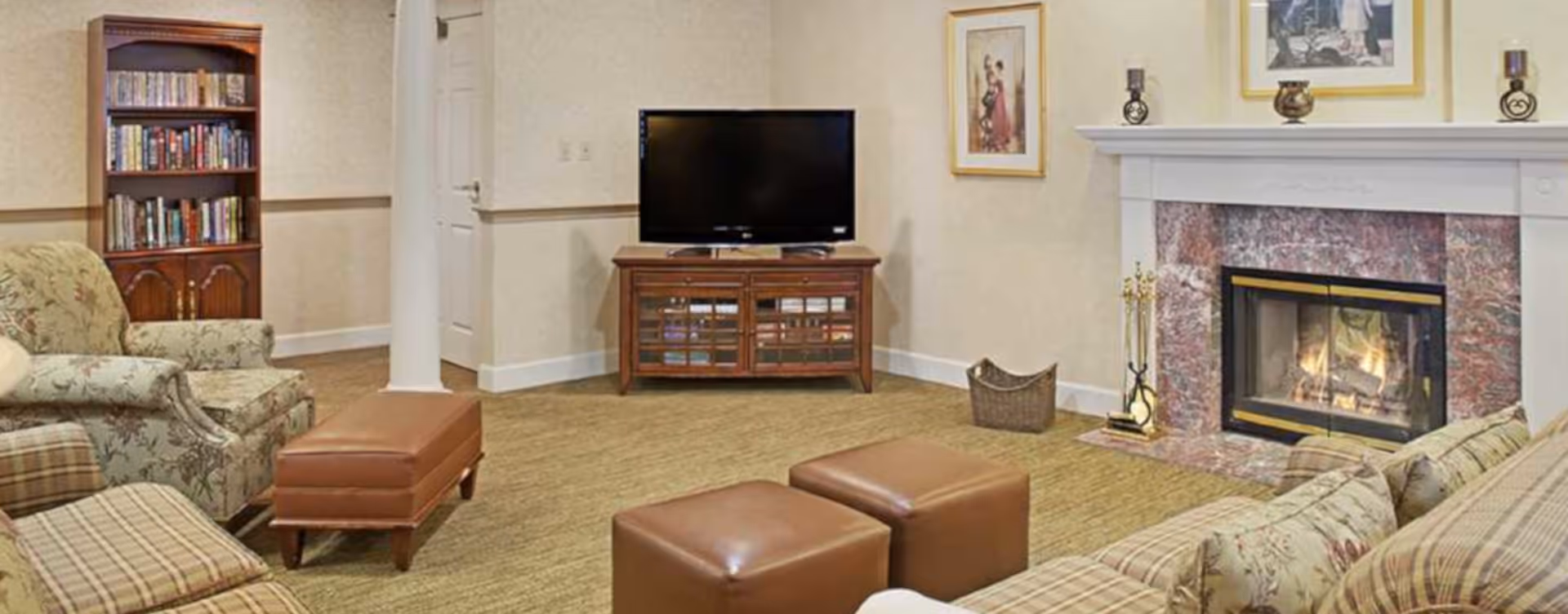 Cozy common living room with upholstered chairs and ottomans, a TV and bookshelf, and a lit fireplace.